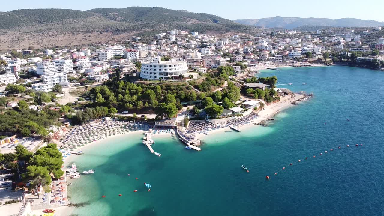 Ksamil, Arnavutluk - Otelleri, şezlongları, plajı, denizi ve su oyun alanıyla popüler bir tatil beldesinin geriye doğru havadan çekimi.