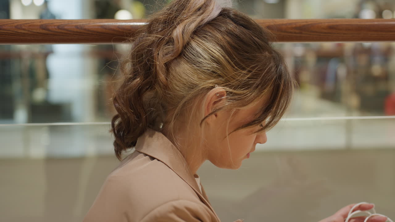 난간 근처에서 스마트폰에 집중하고 있는 어린 소녀, 쇼핑몰 난간에서 휴대폰을 사용하고 있는 흰색 십 대 소녀, 쇼핑몰 난간에서 휴대폰에 집중하고 있는 땋은 머리를 한 십 대 소녀