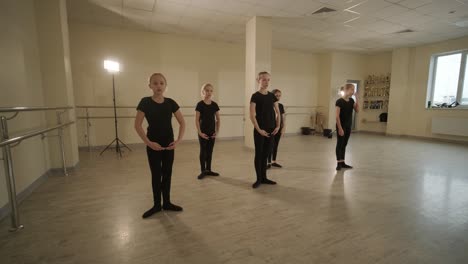 a group of young ballet students in black dancewear practicing positions in a spacious ballet studio with wooden flooring and wall-mounted barres. focused expressions and synchronized movements.