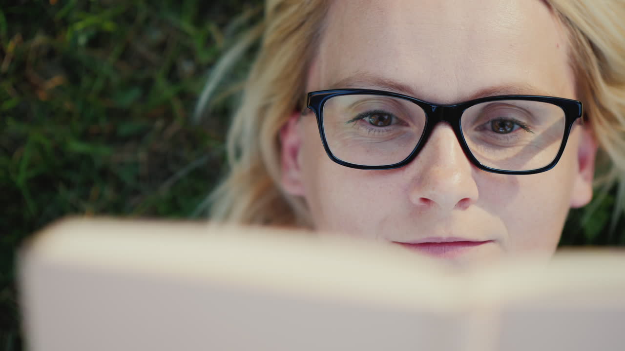 retrato de una mujer joven con gafas tumbada en un césped en el parque y leyendo un libro de cerca 4k vide