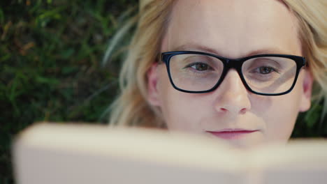 Retrato-De-Una-Mujer-Joven-Con-Gafas-Tumbado-En-Un-Césped-En-El-Parque-Y-Leyendo-Un-Primer-Plano-Del-Libro-4k-Vide