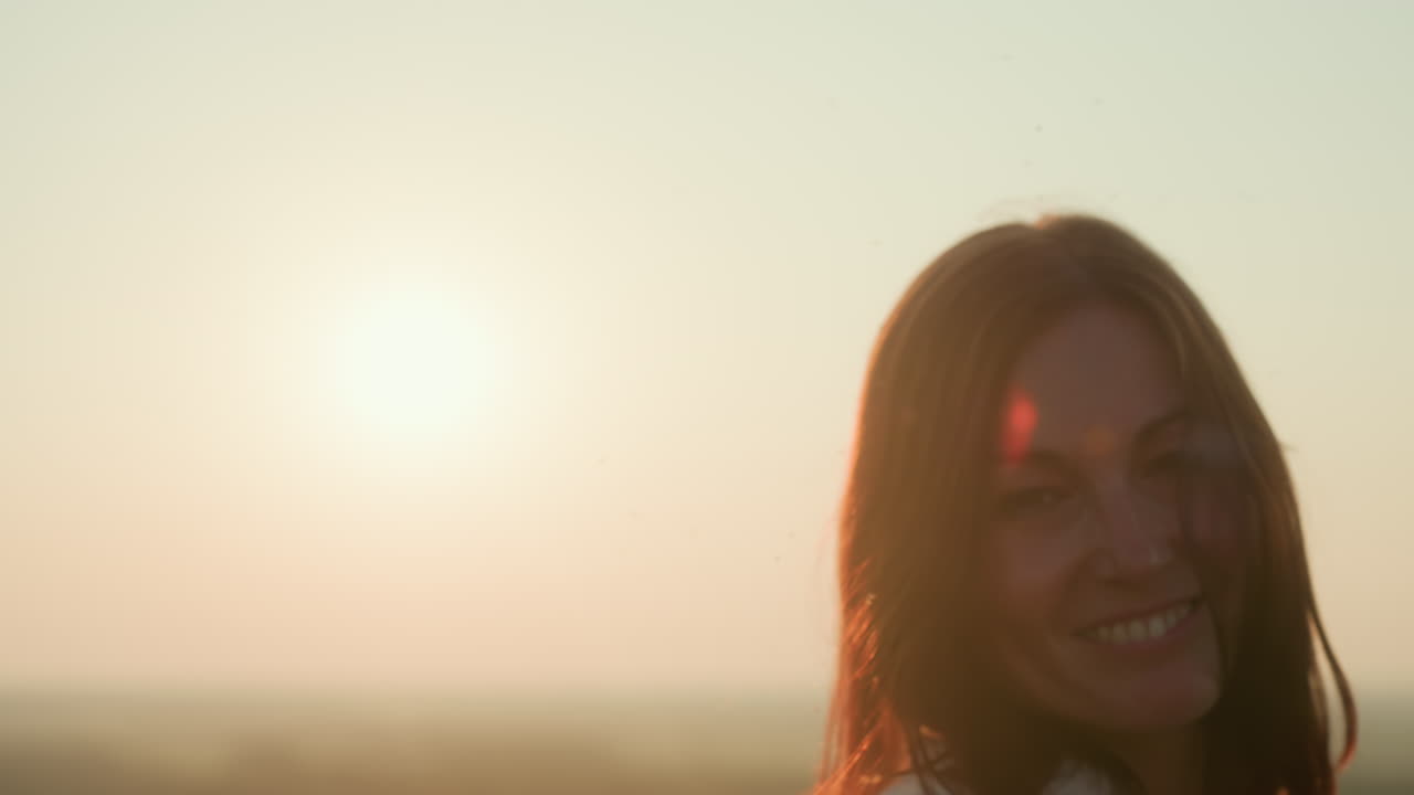 Close up silhouette of young woman twirling joyfully under sun flare as hair flows in breeze across grassy hill top with distant river valley blurred in warm glow creating sense of freedom and bliss