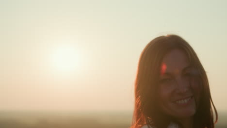 close up silhouette of young woman twirling joyfully under sun flare as hair flows in breeze across grassy hill top with distant river valley blurred in warm glow creating sense of freedom and bliss