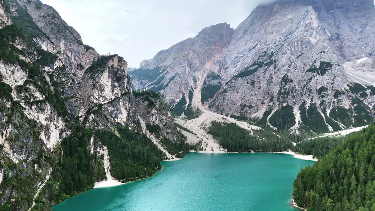 Veduta aerea con drone delle acque turchesi del Lago di Braies circondato da una fitta foresta alpina e imponenti montagne calcaree nelle Dolomiti, Italia