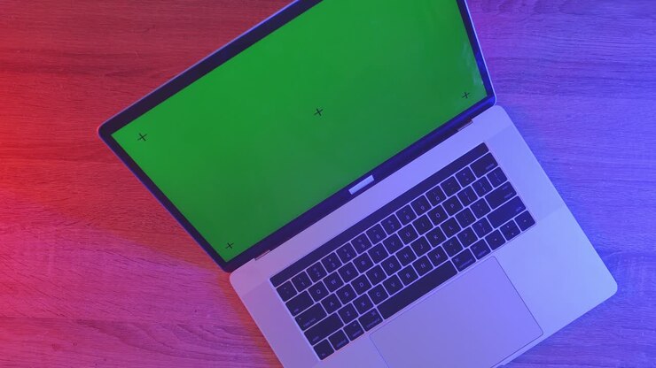 Draufsicht auf einen Laptop-Computer mit Mockup-Greenscreen-Display auf einem Holzschreibtisch, daneben ein Notizbuch mit Stiften, Brille und einem Glas Wasser. Langsames Herauszoomen, Nahaufnahme.
