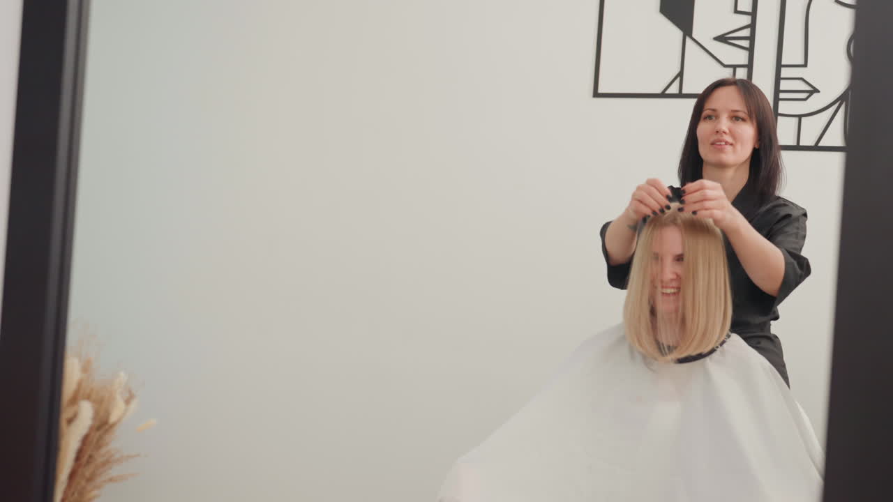 Blonde Kundin sitzt mit Umhang im Salon, Stylist passt den Umhang an und begutachtet das feuchte Haar, sanftes Kämmen und leichtes Schneiden führen zum abschließenden Styling. Minimalistisches Studio mit neutralen Wänden, weiches natürliches Licht