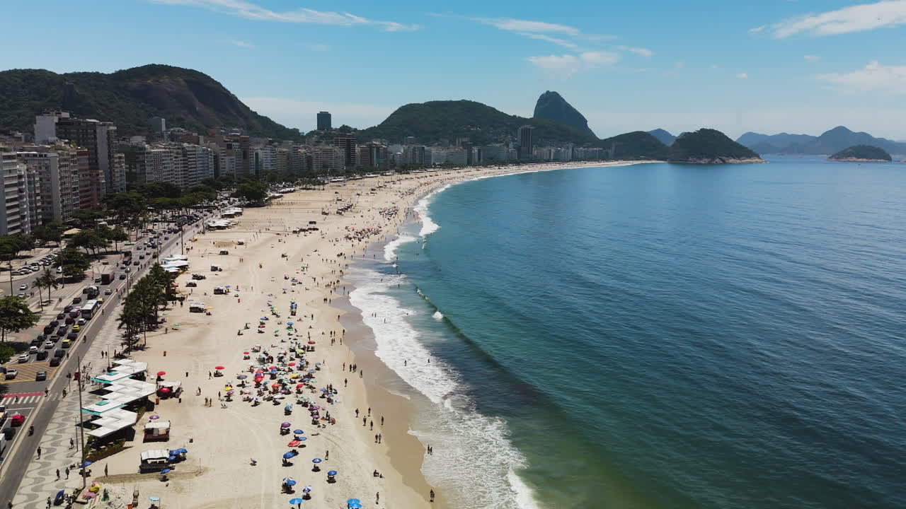 Aerial flyover descent of Copacabana Beach backed by Sugarloaf Mountain on sunny Rio day