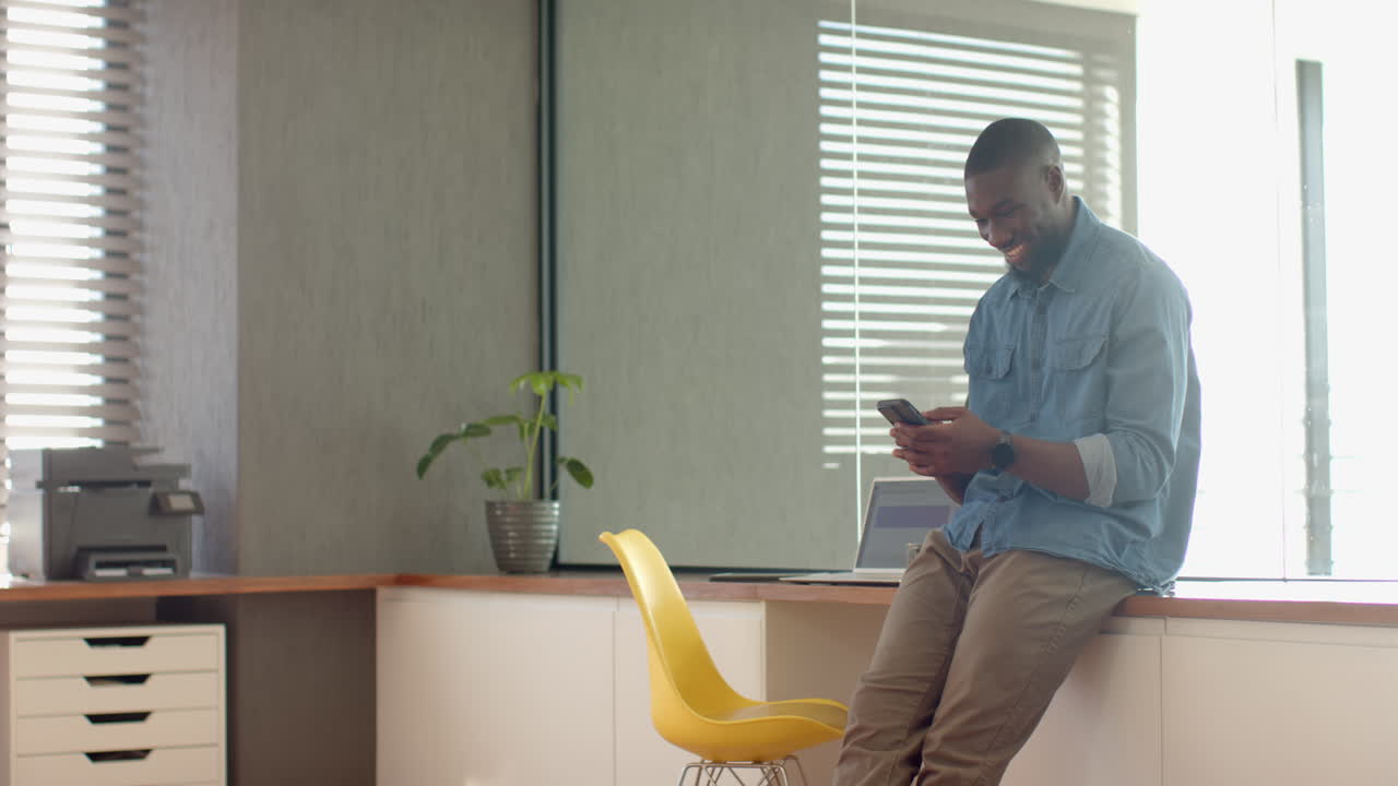 Using smartphone, smiling African American man sitting on desk in modern home office