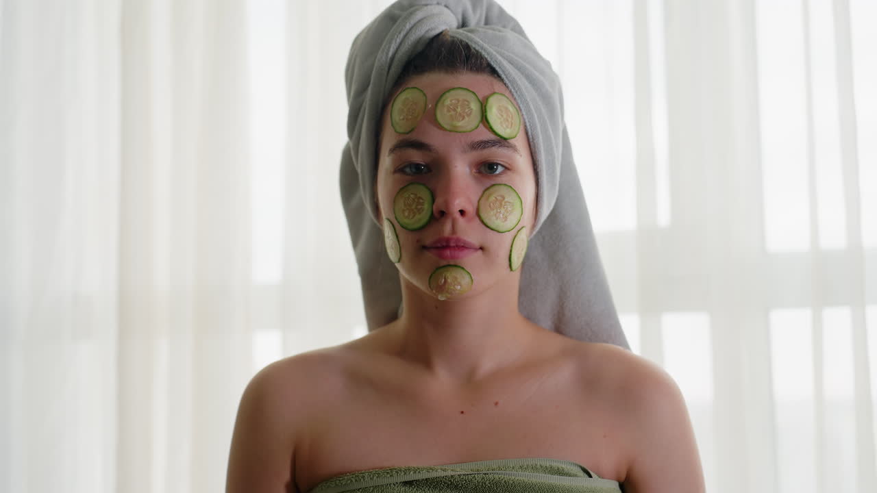 Portrait view of young woman with cucumber slices on face, standing with focused expression, wrapped in towel, enjoying refreshing skincare and calming wellness ritual under soft natural light