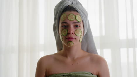 portrait view of young woman with cucumber slices on face, standing with focused expression, wrapped in towel, enjoying refreshing skincare and calming wellness ritual under soft natural light