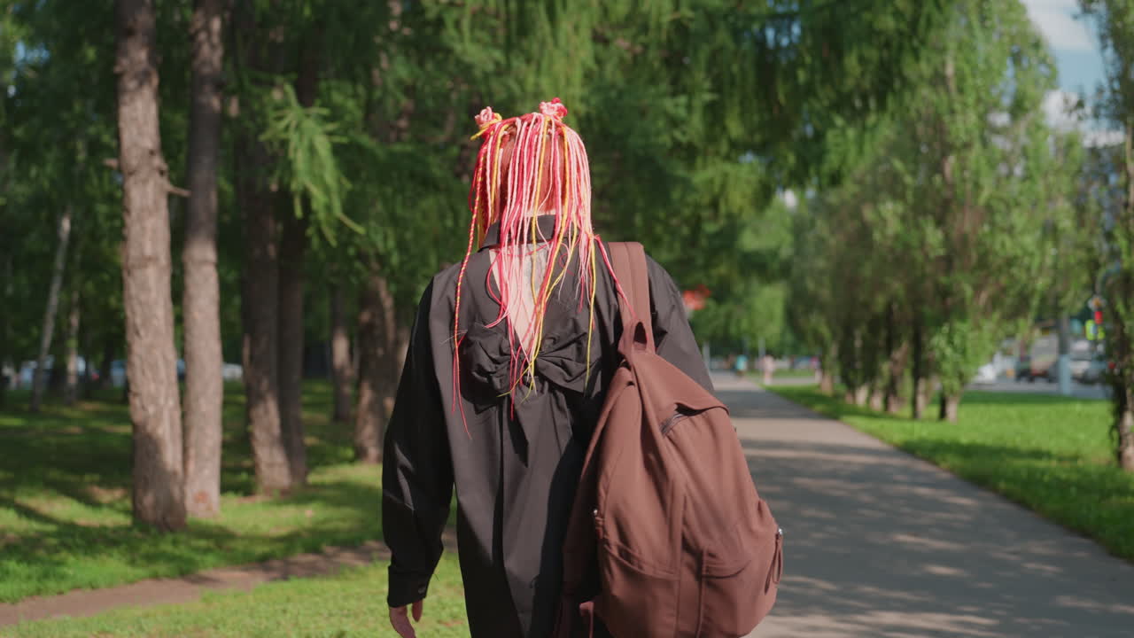 Mujer paseando de forma contemplativa, paseo por el parque con rastas coloridas, trotando por el parque con una mochila grande, mujer reflexiva en un camino sombreado del parque llevando un bolso de gran tamaño y rastas llamativas