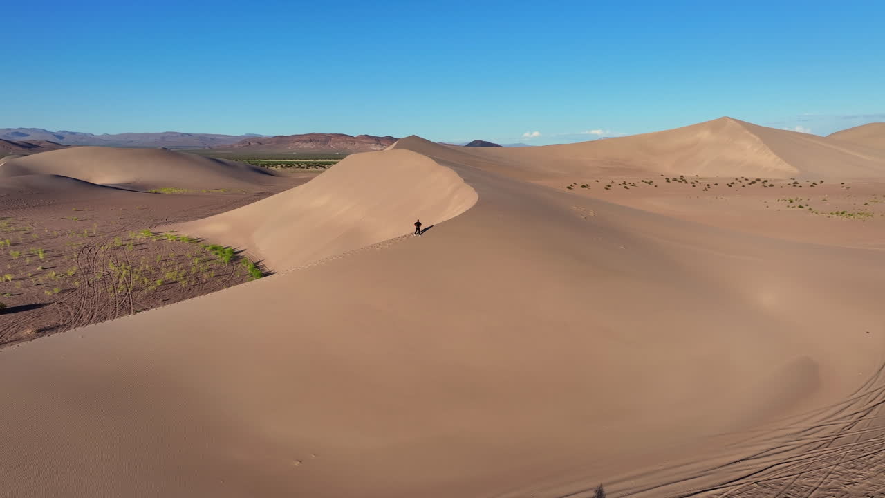 A man climbs up a desert dune in the sunshine. The shot is from a drone circling above.
