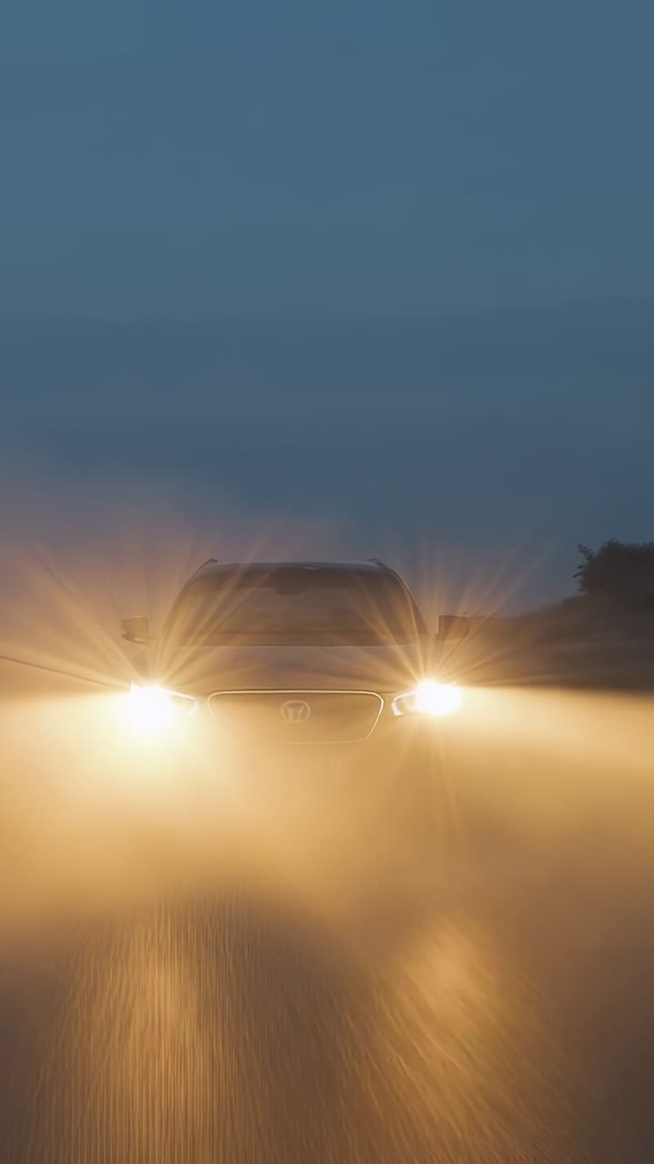 Vídeo vertical: neblina se dissipando e revelando um veículo se aproximando em uma estrada molhada, com faróis brilhantes iluminando as árvores