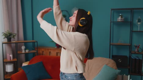 overjoyed young woman in wireless headphones dancing, singing on cozy couch in living room at home