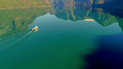 Magical-aerial-shot-of-a-ferry-making-ripples-in-the-water-of-perfectly-still-lake-reflecting-the-mountains-and-the-sky-on-a-sunny-day