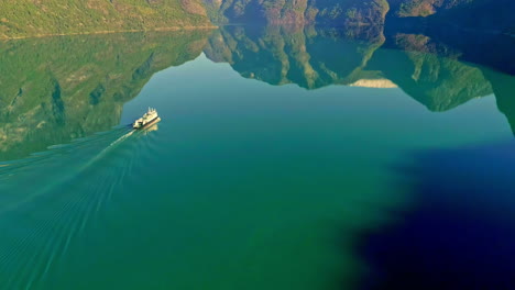 toma aérea mágica de un ferry que hace ondas en el agua de un lago perfectamente quieto que refleja las montañas y el cielo en un día soleado