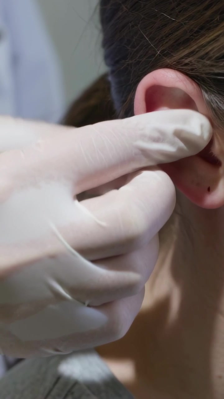 Video verticale: Mano guantata che si avvicina e solleva l'orecchio destro in una clinica, controllando un piercing, con spazio per il testo