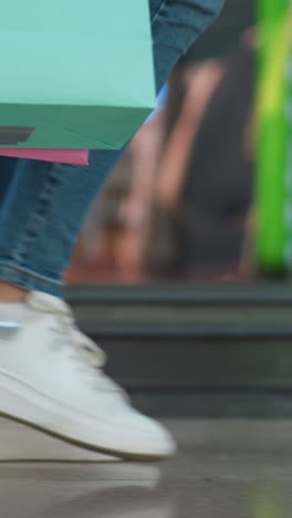vista lateral de una mujer en vaqueros y zapatillas de deporte de lona que lleva una bolsa de compras de menta y rosa, caminando en un centro comercial bien iluminado con su reflejo visible en el vidrio