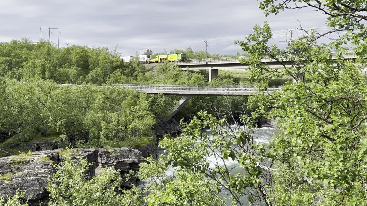 A long cargo train passing over a bridge with a white water river underneath in northern Sweden. Surrounded by nature on a cloudy day. Static shot