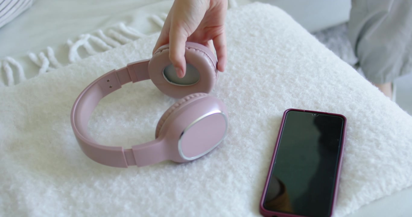 Donna seduta a casa, con la mano destra che si allunga per prendere delle cuffie rosa per la musica da un plaid, accanto a un telefono viola.