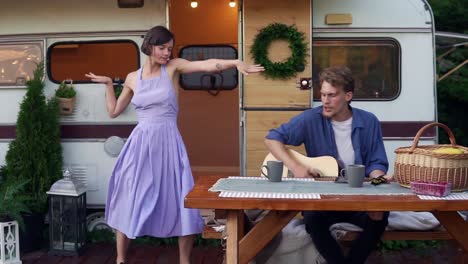 Funny-girl-in-blue-summer-dress-girl-dancing-while-man-playing-guitar-sitting-at-the-wooden-table-in-front-trailer,-singing-a-song-loudly.-Vacation,-holidays,-trailer-trip