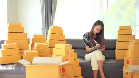 young asian woman, owner of online shop, making list of inventory and orders while sitting on sofa with product in boxes, full frame slow motion