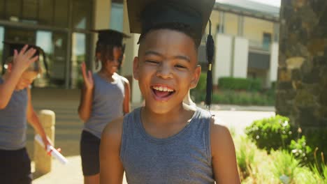 video de un feliz niño afroamericano con sombrero de graduación frente a la escuela