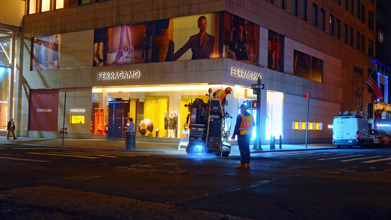 New York, USA, 16 aprile 2026: Macchinari per la costruzione stradale al lavoro sulla Fifth Avenue di notte. Fresatrice stradale e operai al lavoro di notte davanti a una vetrina di moda di lusso.