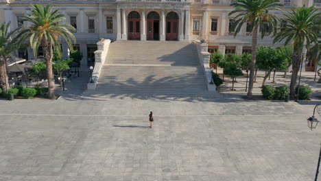 Drone-shot-of-a-woman-walking-towards-the-city-hall-of-Ermoupoli-in-Syros,-Greece-during-sunrise