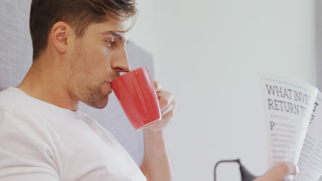 Young-man-sitting-on-bed-having-coffee-while-reading-newspaper-4K-4k