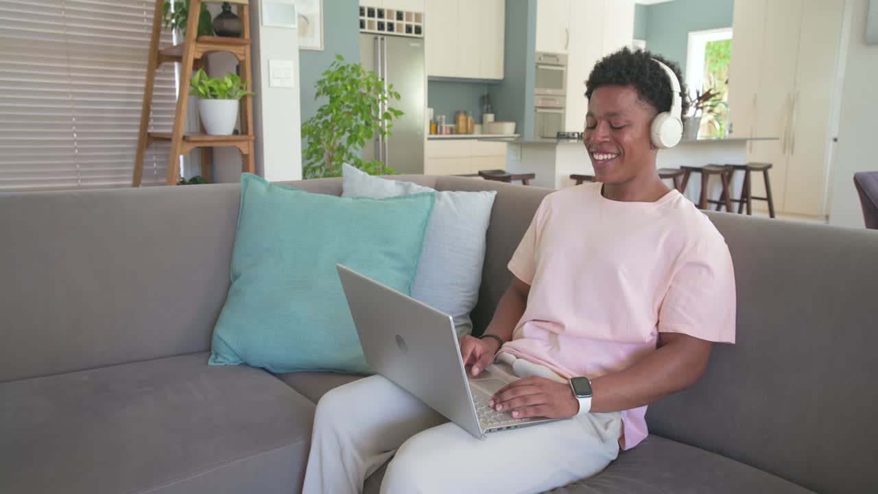 African-American male student reacting to laptop audio, typing on laptop on gray couch with headset