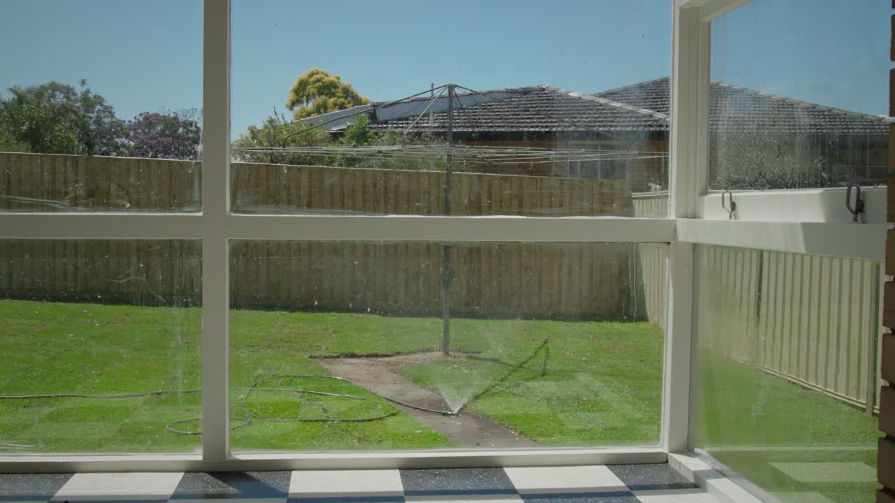 Vy inifrån ett bostadshus som tittar ut genom ett lätt smutsigt fönster mot en förortsbakgård, där en vattenspridare vattnar en grön gräsmatta. Med en klassisk australisk hill hoist-torkvinda.