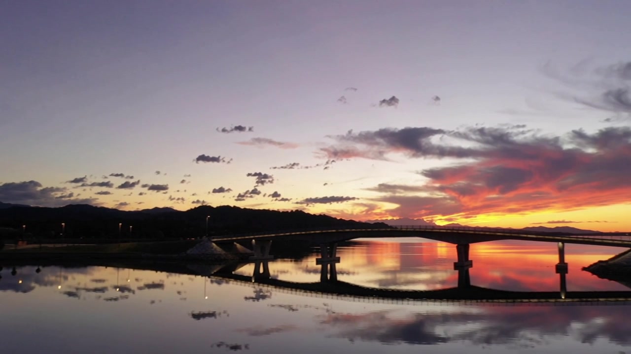 Il sole che tramonta, il cielo al tramonto che si intensifica proiettando colori sulla riva, riflettendo un ponte e l'acqua calma.