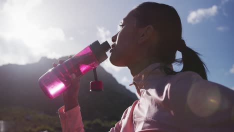 Mujer-Afroamericana-Bebiendo-Agua-De-Una-Botella-Mientras-Camina-Por-Las-Montañas