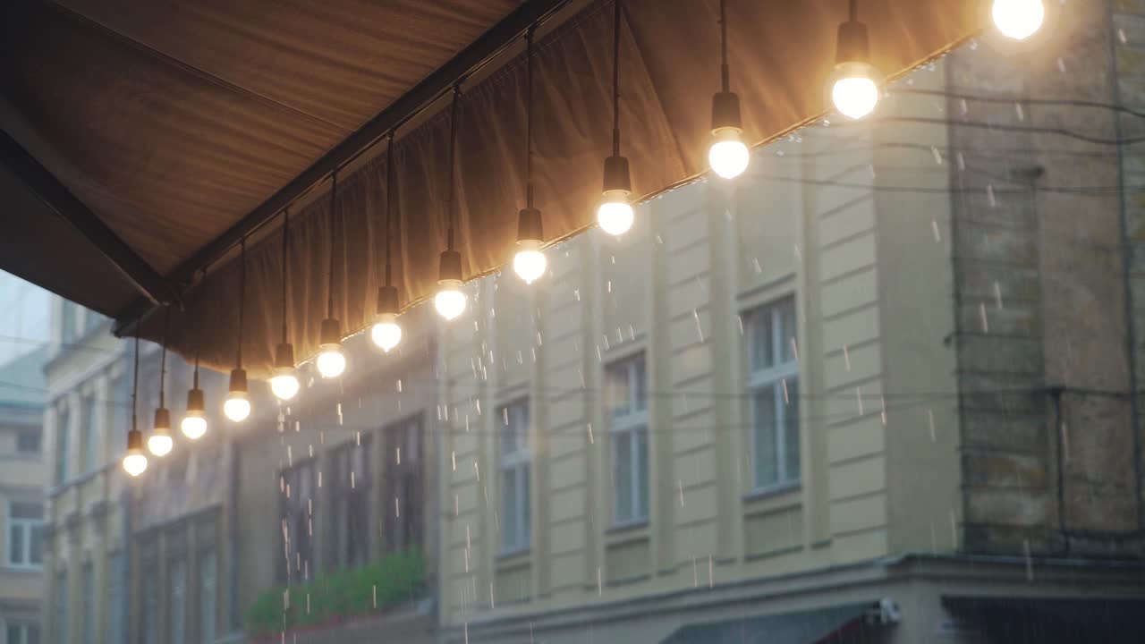 view on a row of burning light bulbs from the roof of a restaurant on the background of raindrops from the street. It's raining