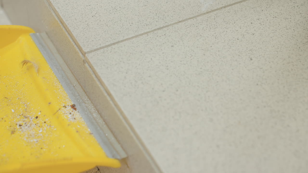High angle view shows dirt, dust, crumbs on tiled floor while hand with broom stick sweeps debris toward pan beside washer, cleanup, bristles push along grout during maintenance in laundry room