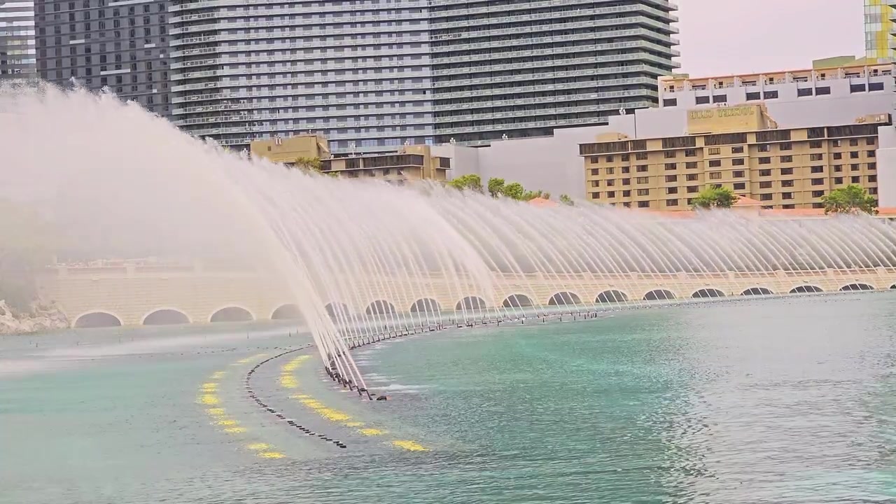 Toma amplia de chorros de fuente coreografiados sobre un pintoresco lago de un complejo turístico, con agua color cerceta, niebla, arquitectura de piedra arqueada, hoteles de gran altura, vegetación y un ambiente vacacional soleado para viajar