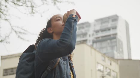 Happy-biracial-woman-in-city,-wearing-backpack-and-drinking-takeaway-coffee