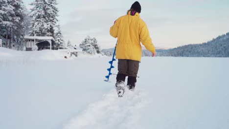 back view of a man walking in thick snow with ice auger, going to drill hole for ice fishing in winter