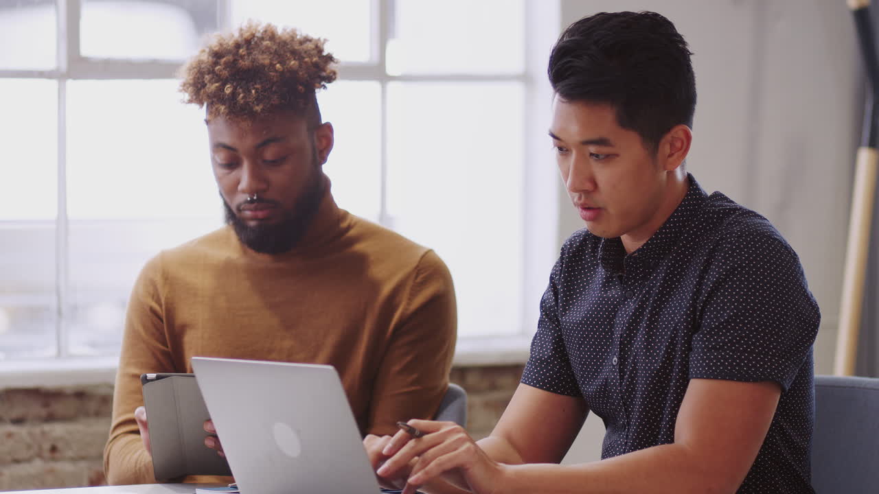Two male creatives using a laptop and tablet in a meeting with an unseen colleague, close up