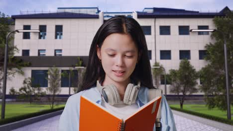 close up of asian teen girl student with a backpack holding and reading a book while standing in front of a school building