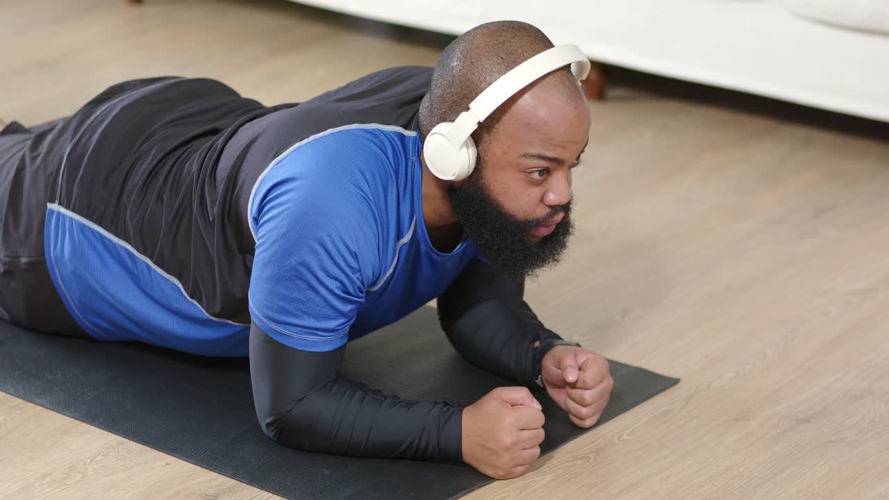 Afrikaanse man in sportkleding doet een onderarmplank op een mat thuis, volgt een audiocue en traint zijn core.
