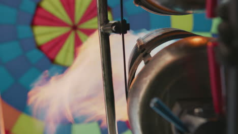 close up metal burner releasing intense flame into colorful hot air balloon canopy with vibrant radial pattern of red and yellow inside blue panels under bright daylight sky