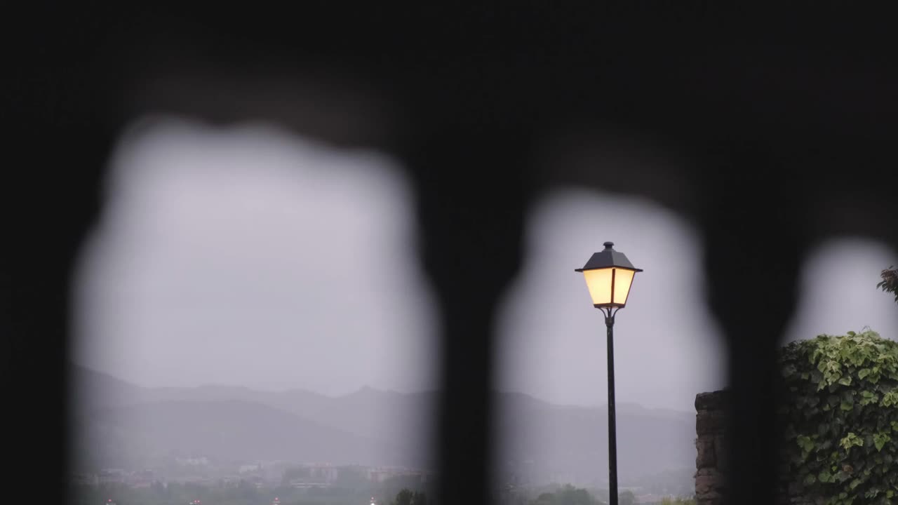 Street lamp glowing at dusk framed by iron railing with mountains and town visible in the background in Hondarribia, Spain