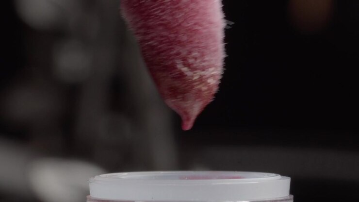 Scatto macro che mostra un applicatore per trucco rivestito di rosso inserito in una bottiglia di cosmetico durante la preparazione di un prodotto di bellezza, con texture dettagliata e messa a fuoco ravvicinata.