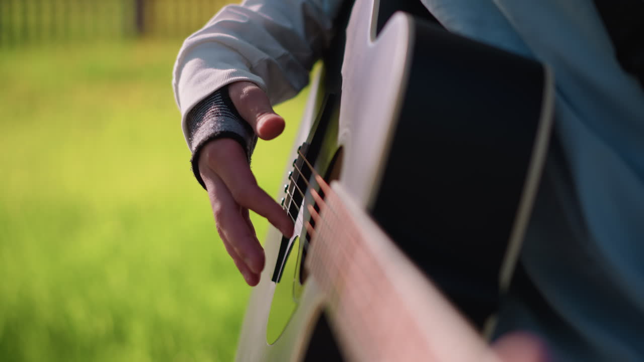 Un chitarrista caucasico che pizzica una chitarra acustica all'aperto, soffusa luce mattutina, prato verde sullo sfondo, inquadratura ravvicinata sulla mano destra, delicati movimenti delle dita sulle corde, manica di jeans e polsino in pelle