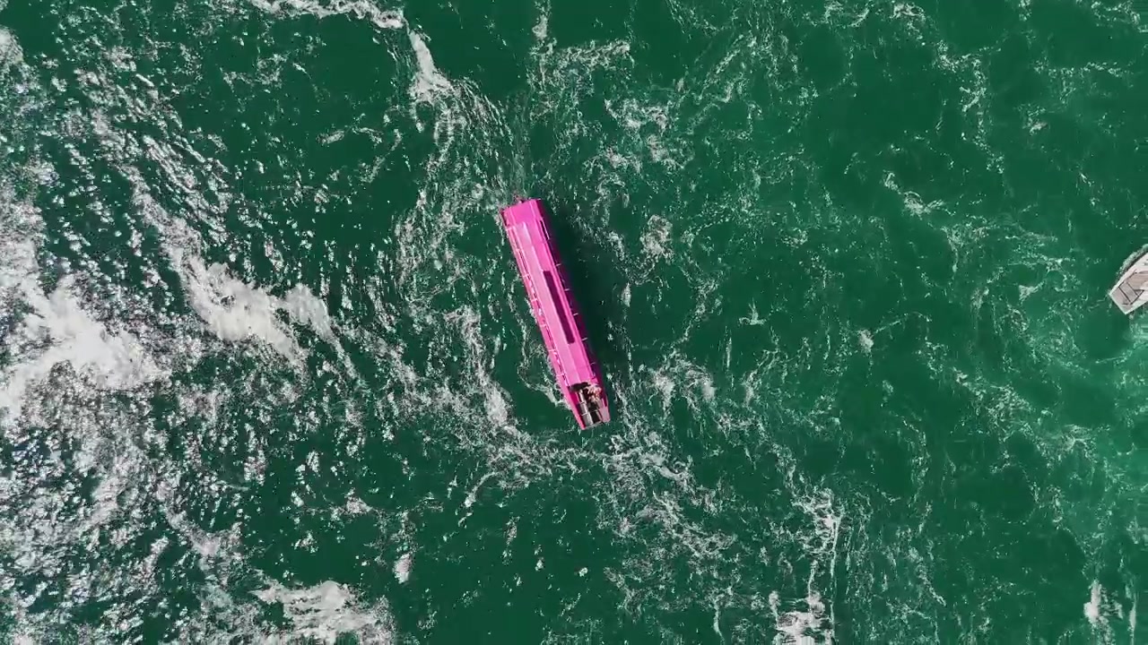 Top-down drone view of a bright pink passenger boat crossing turbulent turquoise water in Schaffhausen, Switzerland, surrounded by swirling currents and foamy patterns.
