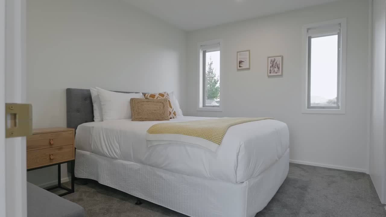 Acercamiento lento a un espacio para dormir limpio y moderno con cama blanca, mesita de noche de madera, manta decorativa amarilla y arte de pared enmarcado con ventanas que muestran árboles al exterior