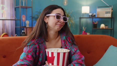 young woman sits on sofa eating popcorn watching interesting tv serial sport game online at home