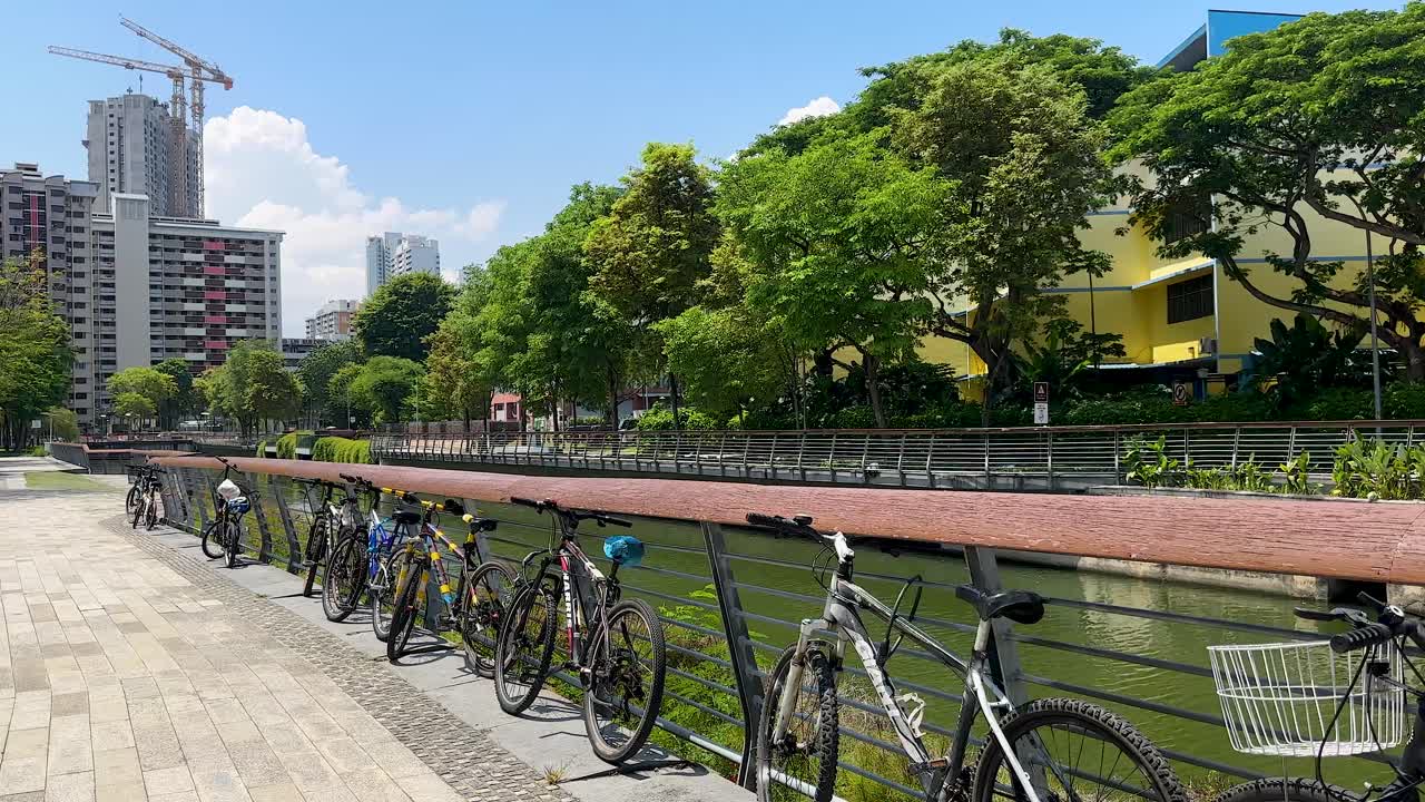 Daytime camera pan past parked bicycles on canal bridge, modern cityscape and greenery visible